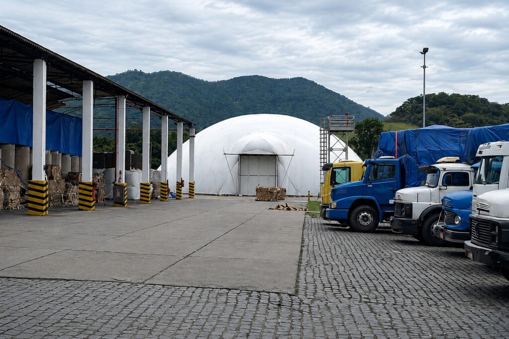 Ao fundo um galpão e ao lado caminhões e uma cobertura