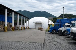 Ao fundo um galpão e ao lado caminhões e uma cobertura