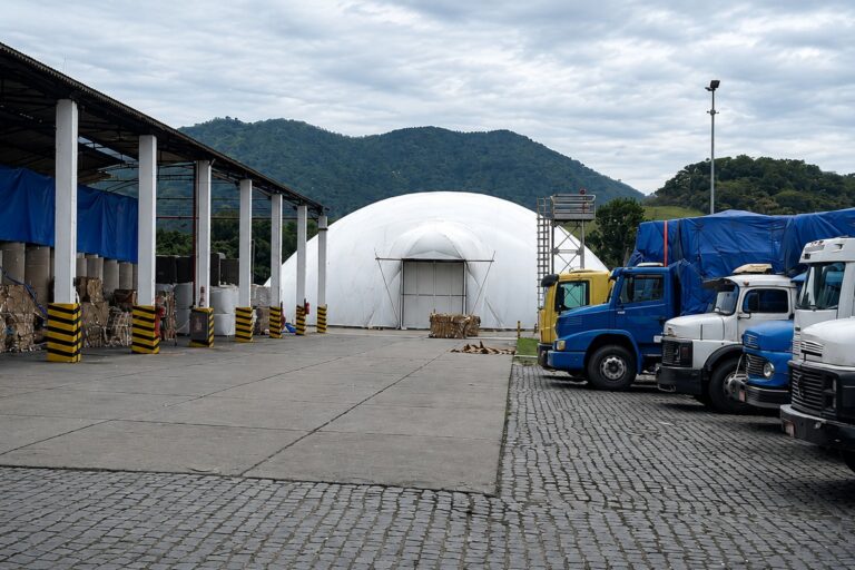 Ao fundo um galpão e ao lado caminhões e uma cobertura